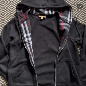Svart hoodiejacka med rutigt foder - Svart hoodiejacka med dragkedja och stora fickor framtill. Insidan av huvan har ett rutigt mönster i svart, vitt, grått och rött som ger en snygg kontrast. Jackan har snörning i huvan och är perfekt för chill dagar.