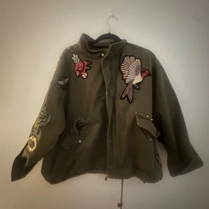 Olivgrön parkajacka med broderier - Säljer en olivgrön parkajacka med coola broderier av fågel, ros och orm. Jackan har dragsko nertill, stora fickor och knappar framtill. Oversized passform och unika detaljer som verkligen sticker ut. Perfekt för dig som vill ha något extra i garderoben.