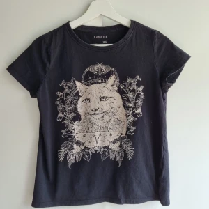 Svart t-shirt med kattmotiv från Medicine - Svart t-shirt från Medicine i bomull med korta ärmar och rund hals. Framsidan har ett detaljerat tryck med naturtema, bland annat en katt, nattfjäril och växter i beige. Perfekt skick.