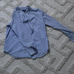 Randig skjorta från Ralph Lauren - Snygg blå- och vitrandig skjorta från Ralph Lauren med klassisk krage och knäppning framtill. Skjortan har lång ärm och ikonisk broderad logga på bröstet. Tillverkad i mjuk bomull som känns skön mot huden. Perfekt för dig som gillar stilren och tidlös look. ( i XS )