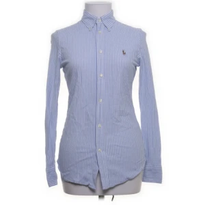 Blårandig skjorta från Ralph Lauren - Klassisk blårandig skjorta från Ralph Lauren med broderad logga på bröstet. Skjortan har lång ärm, knappar hela vägen och krage med knappar. Tillverkad i mjuk bomull och har en slim passform som sitter snyggt. Perfekt för en clean och stilren look.