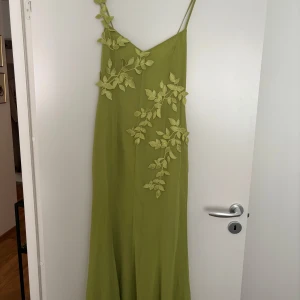 Grön långklänning med broderade blad NA-KD - Superfin grön långklänning från NA-KD med smala axelband och v-ringning. Klänningen har broderade bladapplikationer framtill och på axelbandet, samt knytning i ryggen. Materialet är lätt och flowy, perfekt för sommarens alla tillfällen.