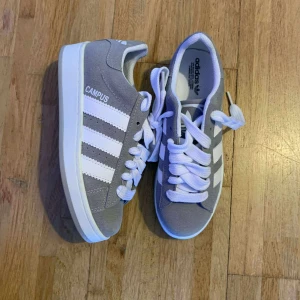 Grå Adidas Campus sneakers - Snygga grå Adidas Campus sneakers med klassiska vita ränder och tjocka vita snören. Ovandel i mocka och platt vit sula. Perfekta för en clean och sportig stil.
