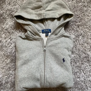 Grå hoodie från Polo Ralph Lauren - Snygg grå hoodie från Polo Ralph Lauren med dragkedja och klassisk broderad logga på bröstet. Tröjan har huva och är tillverkad i mjuk bomullsmix med inslag av polyester. Perfekt för chill dagar och enkel att matcha med jeans eller joggers.                            Storlek 14-16 (ca S/XS).                                                Mått 📏 axelbred 43 cm längd 55 cm ärm 55 cm.            Hör av dig vid frågor eller för fler bilder! 
