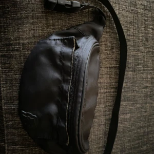 Svart magväska från ZARA med röd dragkedja - Säljer en svart magväska från ZARA i syntetmaterial. Väskan har en röd dragkedja som ger en cool kontrast och ett justerbart band. Perfekt för att bära mobilen, plånboken och andra smågrejer när du är på språng.