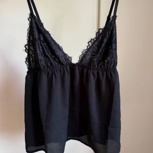 Svart spetslinnetopp med tunna band - Supersnygg svart linnetopp med tunna axelband och spetsdetaljer upptill. Toppen har en luftig, lätt genomskinlig nederdel och justerbara band för perfekt passform. Perfekt att styla med jeans eller kjol för en trendig look.