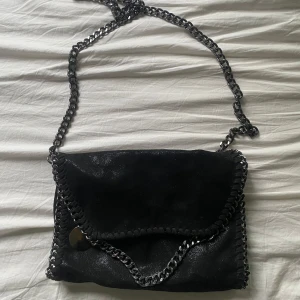 Svart axelväska med kedjedetaljer - Cool svart axelväska med glansig yta och grova kedjor längs kanterna och axelremmen. Väskan har ett lock och insidan är fodrad med leopardmönster samt har dragkedjefack. Köpt på amazon och har lite tecken på användning men inte något som märks av