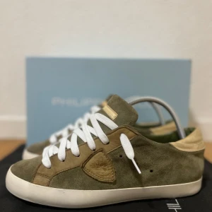 Olivgröna Philippe Model sneakers i mocka - Säljer ett par olivgröna Philippe Model sneakers i mocka med vita snören och beige detaljer. Skorna har en vit platt sula och rund tå. Klassisk design med märkets logga på sidan. Perfekta för dig som gillar stilrena och bekväma sneakers. Storlek 40 passar också stl 41. Bra skick! 