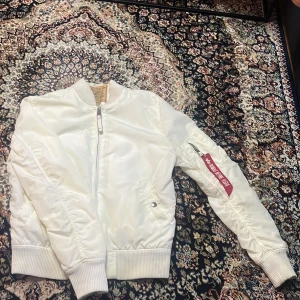 Vit bomberjacka Alpha Industries M - Snygg vit bomberjacka från Alpha Industries i storlek medium. Jackan har klassisk passform, ribbade muddar och dragkedja framtill. På ärmen finns en ficka med den ikoniska 'Remove Before Flight'-taggen. Tillverkad i glansig polyester för en clean look.