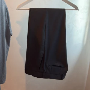 Svarta kostymbyxor med uppvik - Snygga svarta kostymbyxor med klassisk passform och uppvikta benslut. Byxorna har en stilren look med diskreta fickor bak och är perfekta för en dressad outfit. Materialet känns mjukt och har en lätt struktur.