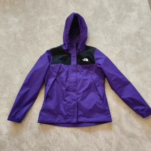 Lila vindjacka från The North Face - Snygg lila vindjacka från The North Face med svarta axelpartier och huva. Jackan är tillverkad i slitstark polyester med DryVent-teknologi som håller dig torr. Perfekt för blåsiga dagar och har dragkedja framtill samt elastiska ärmslut. 