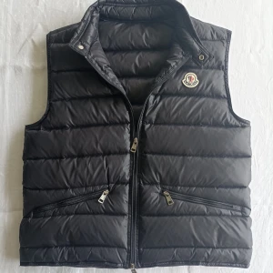 Svart dunväst från Moncler - Snygg svart dunväst från Moncler med klassisk quiltad design och hög krage. Västen har två dragkedjefickor framtill och stängs med dragkedja. Ikonisk Moncler-logga på bröstet ger en exklusiv känsla. Perfekt att slänga över en hoodie eller tröja.NFC scan finns.