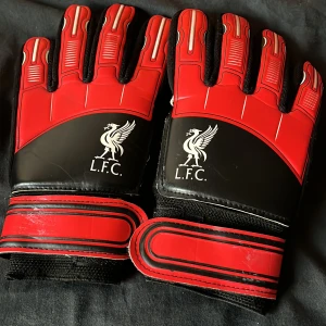 Liverpool FC målvaktshandskar röd/svart - Snygga målvaktshandskar i rött och svart med Liverpool FC-logga på ovansidan. Handskarna har breda kardborreband vid handleden och vita insidor för bra grepp. Perfekta för dig som vill visa ditt LFC-stöd på fotbollsplanen.