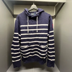 Randig hoodie från Polo Ralph Lauren - Mörkblå och vit randig hoodie från Polo Ralph Lauren med huva och snörning. Tröjan har känguruficka framtill och är tillverkad i bomullsmix. Perfekt för en chill och avslappnad stil.