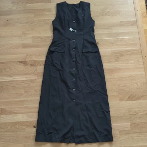 Svart långklänning med knappar - Stilren svart långklänning med v-ringning och knäppning hela vägen framtill. Lite kostym vibe. Klänningen är ärmlös och har två fickor med lock framtill samt snygga knappdetaljer i ryggen. Perfekt för dig som gillar klassisk och enkel stil.