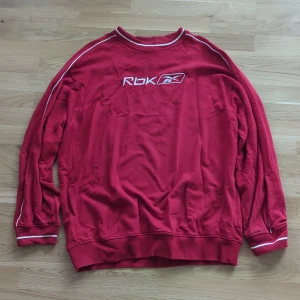 Röd crewneck från Reebok XXL - Röd crewneck från Reebok med vit RBK-logga broderad på bröstet. Tröjan har vita ränder längs ärmarna och runt kragen samt muddar vid ärmslut och nederkant. Tillverkad i mjuk bomull, perfekt för en sportig och avslappnad stil. Enligt loggan är den från 2006-2008.