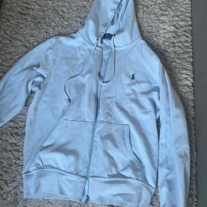 Ljusblå hoodie från Polo Ralph Lauren - Snygg ljusblå hoodie från Polo Ralph Lauren med dragkedja, huva med snörning och klassisk broderad logga på bröstet. Tröjan har känguruficka och ribbade muddar. Perfekt för en chill och stilren look.om man är S så funkar den också.