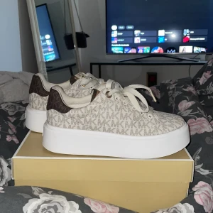 Beige sneakers från Michael Kors - Snygga sneakers från Michael Kors med beige och brunt MK-mönster, vita snören och kraftig vit platåsula. Skorna har detaljer i brunt och ljusbrunt konstläder vid häl och plös, samt guldfärgade öljetter. Perfekta för dig som gillar statement-sneakers.