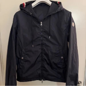 Mörkblå vindjacka från Moncler  - Säljer en Navyblue windbreaker från Moncler med huva och dragkedja framtill. Jackan har långa ärmar, två fickor med knappar och diskret Moncler-logga på ärmen. Riktigt najs nu till sommarn och hösten sen. Snygga detaljer med snörning i huvan. Jackan är i storlek 3 vilket motsvarar M,L. Kan gå ner i pris via en snabb smidig affär✌🏽