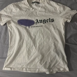 Vit Palm Angels t-shirt Shanghai - Vit t-shirt från Palm Angels med tryck framtill där det står 'Angels Shanghai' och en lila spray paint-effekt över loggan. Den är i bra skick behöver bara strykas ,Klassisk rund hals och korta ärmar. Skön passform och tillverkad i mjuk bomull, perfekt för en clean streetwear-look.