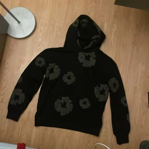 Svart hoodie med blommigt mönster Denim Tears - Säljer en svart hoodie från Denim Tears med gråa blommiga tryck över hela tröjan. Tröjan har huva och är långärmad, perfekt för dig som gillar streetwear med unik design. Tillverkad i USA och i mjukt bomullsmaterial.