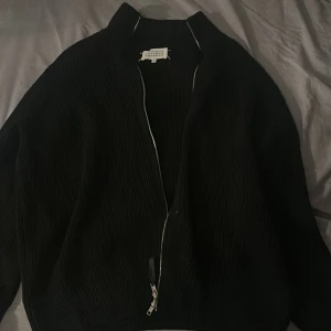Svart cardigan från Maison Margiela - ALDRIG ANVÄND! Svart ribbstickad cardigan jacka från Maison Margiela med dragkedja framtill och klassisk ståkrage. Jackan har diskreta vita stygn på ryggen, vilket är ett signum för märket. Perfekt för dig som gillar minimalistisk och stilren design.