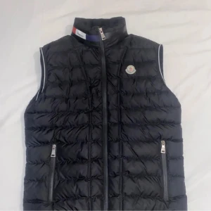 Svart dunväst från Moncler - Säljer en snygg svart dunväst från Moncler med dragkedja och hög krage. Västen har två praktiska fickor med dragkedjor och en stilren logga på bröstet. Perfekt för en sportig look eller som ett extra lager under kyliga dagar.