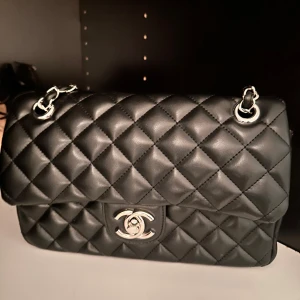 Svart quiltad axelväska  - Säljer en elegant svart quiltad axelväska med silverkedja och klassisk design. Väskan har en praktisk dragkedja inuti och en ficka på baksidan. Perfekt för att ge din outfit en lyxig touch.