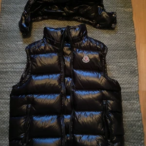 Svart dunväst från Moncler - Säljer en snygg svart dunväst från Moncler med glansig yta och dragkedja framtill. Västen har en avtagbar huva och två praktiska fickor med dragkedja. Perfekt för kyliga dagar när du vill hålla stilen. 🖤 västen passar som S/M 
