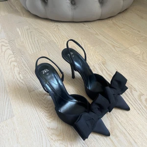 Svarta pumps med rosett från Zara - Eleganta svarta pumps från Zara med spetsig tå och hög klack. Skorna har en stilren design med en stor dekorativ rosett framtill. Klackens höjd är 10 cm. Använda 2 gånger.