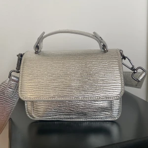 Silver axelväska från HVISK - Snygg silverfärgad axelväska från HVISK med texturerad yta och justerbar axelrem. Väskan har ett större och ett mindre fack och röd insida som ger en fin kontrast. 