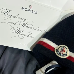 Svart mössa från Moncler - Snygg svart mössa från Moncler med röd och vit randdetalj. Den har en broderad Moncler-logga framtill. Perfekt för att hålla värmen med stil.