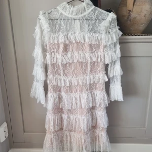 Vit spetsklänning med volanger - Säljer en elegant vit spetsklänning med volanger. Klänningen har långa ärmar och en hög hals, vilket ger en sofistikerad look. Perfekt för speciella tillfällen. 💫