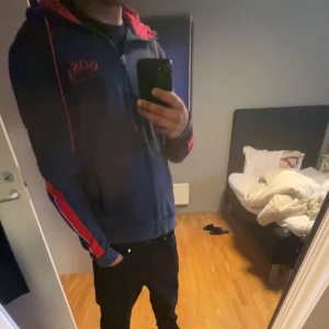 Mörkblå hoodie från Boss - Säljer en mörkblå hoodie från Boss med röda detaljer och dragkedja. Den har en justerbar huva med röda snören och långa ärmar med röda ränder längs sidorna. Perfekt för en avslappnad stil.