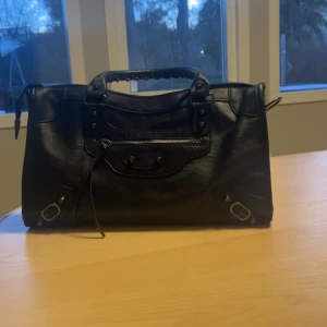 Svart axelväska med dragkedja - Snygg svart axelväska i skinn med praktisk dragkedja och avtagbar axelrem. Väskan har ett stilrent utseende med dekorativa spännen och handtag. Perfekt för att bära dina dagliga nödvändigheter med stil.