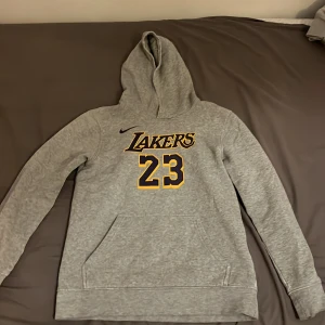 Grå Lakers hoodie från Nike - Säljer en grå hoodie från Nike med Lakers-logga och nummer 23 på framsidan. Baksidan har texten 'James' och nummer 23. Perfekt för basketfans! Hoodien har en klassisk design med långa ärmar och en bekväm huva.