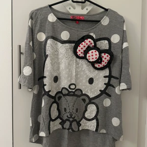 Grå Hello Kitty topp med prickar - Säljer en grå Hello Kitty topp med stora vita prickar och en söt Hello Kitty-figur på framsidan. Toppen har korta ärmar och en lös passform, perfekt för en avslappnad stil. Den har också en rosett med röda prickar som en extra detalj.