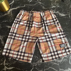 Rutig badshorts från Burberry - Snygga badshorts från Burberry med klassiskt rutigt mönster i beige, svart, vit och röd. De har en elastisk midja med snörning för perfekt passform. Perfekta för sommarens alla badäventyr!