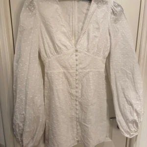 Vit klänning med prickar från Adoore - Säljer en söt vit klänning från Adoore med puffärmar. Klänningen har en v-ringad design och dragkedja i ryggen. Perfekt för vår och sommar!