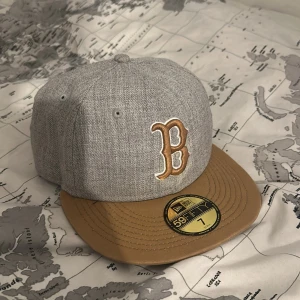 Grå keps från New Era - Snygg grå keps från New Era med en broderad bokstav 'B' i beige framtill. Kepsen har en brun skärm och är i storlek 7. Perfekt för att ge din outfit en cool touch. Pris kan diskuteras också.