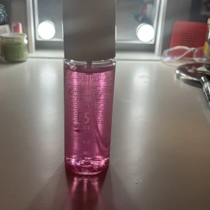 Rosa body mist  - En elegant parfymflaska med rosa vätska och vit kork. Flaskan har en stilren design och är perfekt för den som älskar en fräsch och blommig doft. Passar utmärkt på sminkbordet som en fin detalj.
