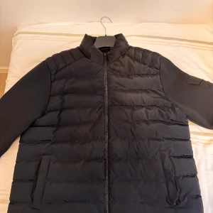 Svart pufferjacka från Moncler - Snygg svart pufferjacka från Moncler med dragkedja och långa ärmar. Jackan har ett stilrent quiltat mönster och en broderad Moncler-logga på ärmen. Perfekt för kyligare dagar.