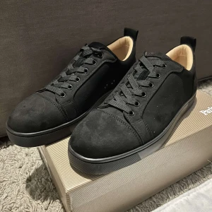 Svarta sneakers från Christian Louboutin - Snygga svarta sneakers i mocka från Christian Louboutin med den ikoniska röda sulan. Skorna har snörning och en rund tå, vilket ger en klassisk look. Perfekta för dig som vill ha stilrena och lyxiga skor.