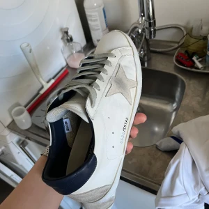 Vita sneakers från Golden Goose - Säljer ett par vita sneakers från Golden Goose med grå stjärndetalj på sidan och grå snörning. Skorna har en mörkblå häl och en klassisk rund tå. Perfekta för en stilren look. Storlek 44 passar 43 dm för mer info og finns