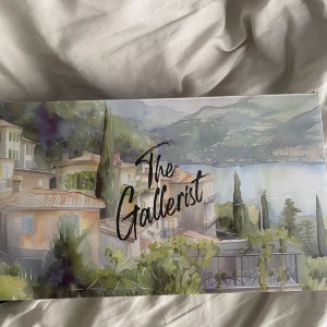 The Gallerist - Säljer brädspelet 'The Gallerist' med ett vackert omslag som visar en pittoresk landskapsmålning med hus och sjö. Perfekt för spelkvällar med vänner och familj. Utforska konstvärlden och bygg ditt eget galleri i detta strategiska spel.