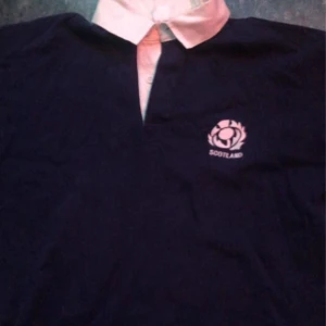 Mörkblå pikétröja med Skottland-emblem - Säljer en mörkblå pikétröja med vit krage och Skottland-emblem på bröstet. Perfekt för sportiga tillfällen eller som en del av en avslappnad outfit.