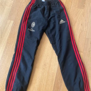 Svarta träningsbyxor från Adidas - Säljer ett par svarta träningsbyxor från Adidas med röda ränder längs sidorna. Byxorna har elastisk midja och logotypen på framsidan. Köpta secondhand. Är i storlek 164 men skulle säga dom passar XS