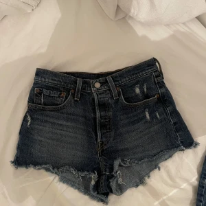 Mörkblå jeansshorts - Säljer ett par snygga blå jeansshorts. De har en klassisk design med både fram- och bakficka samt fransad fåll med två små slitningar framtill. Högmidjade med knapp stängning. 