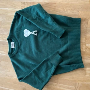 Ami Paris tröja - Säljer en snygg grön tröja från Ami med ett vitt hjärtmönster på bröstet. Tröjan har långa ärmar och ribbade muddar vid ärmslut och nederkant. Perfekt för en stilren look! Extremt skön tröja. Skriv gärna! Pris diskuteras gärna!