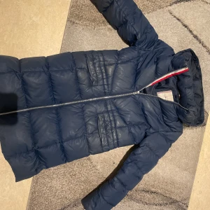 Mörkblå dunjacka från Tommy Hilfiger - Säljer en snygg mörkblå dunjacka från Tommy Hilfiger. Jackan har en dragkedja framtill och en hög krage för extra värme. Den är perfekt för kyliga dagar och har en stilren design med märkets logotyp på ärmen. Ordinarie pris 2,700